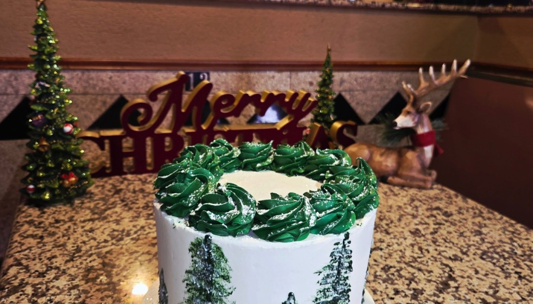 White Christmas cake decorated with a green frosting wreath and pine trees, set against festive decorations.
