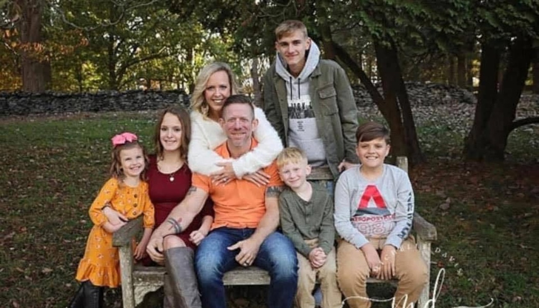 A happy family of seven—parents and five children—smiling together on a wooden bench outdoors.