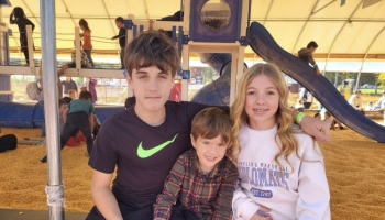 Three smiling siblings, a teenage boy, young boy, and blonde girl, pose together at a covered playground.