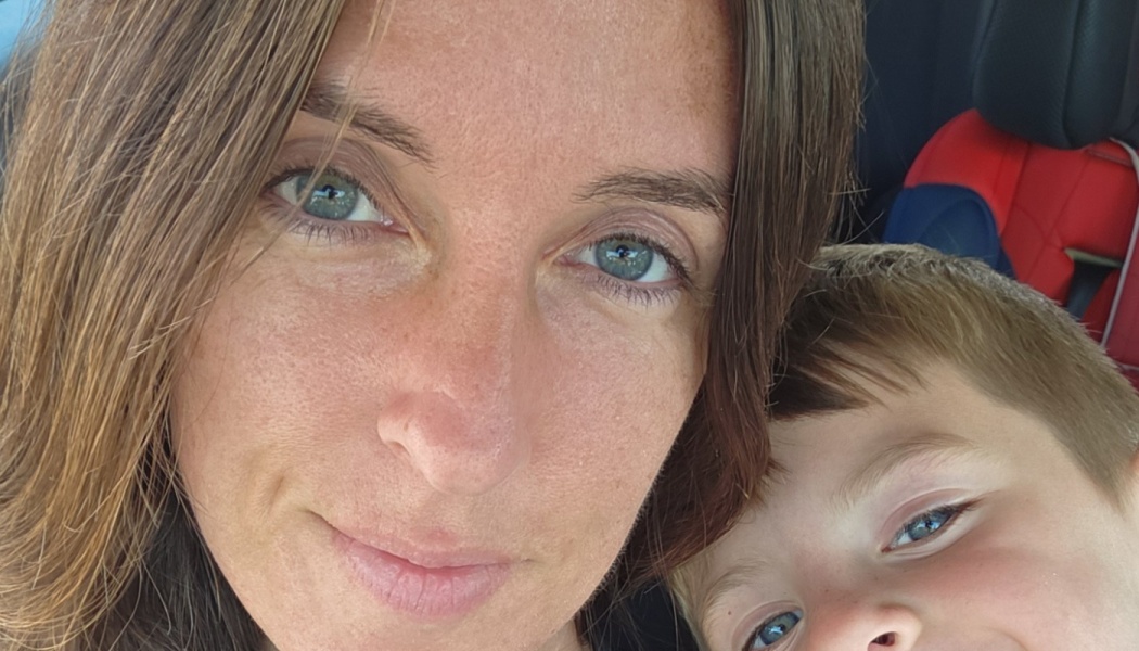 Close-up selfie of a smiling woman with blue eyes alongside her young child inside a car.