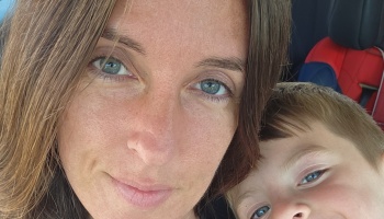 Close-up selfie of a smiling woman with blue eyes alongside her young child inside a car.