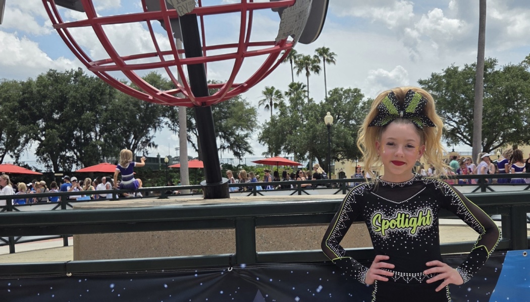 A smiling young cheerleader in a black Spotlight uniform poses outdoors in front of a large red globe sculpture.