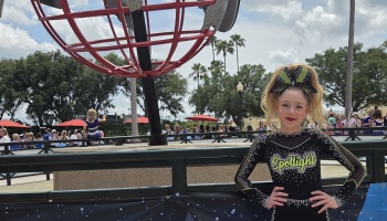 A smiling young cheerleader in a black Spotlight uniform poses outdoors in front of a large red globe sculpture.