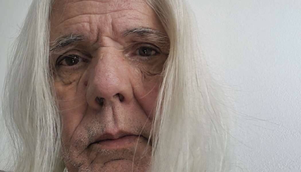 Close-up portrait of an older man with long white hair and brown eyes looking directly at the camera.
