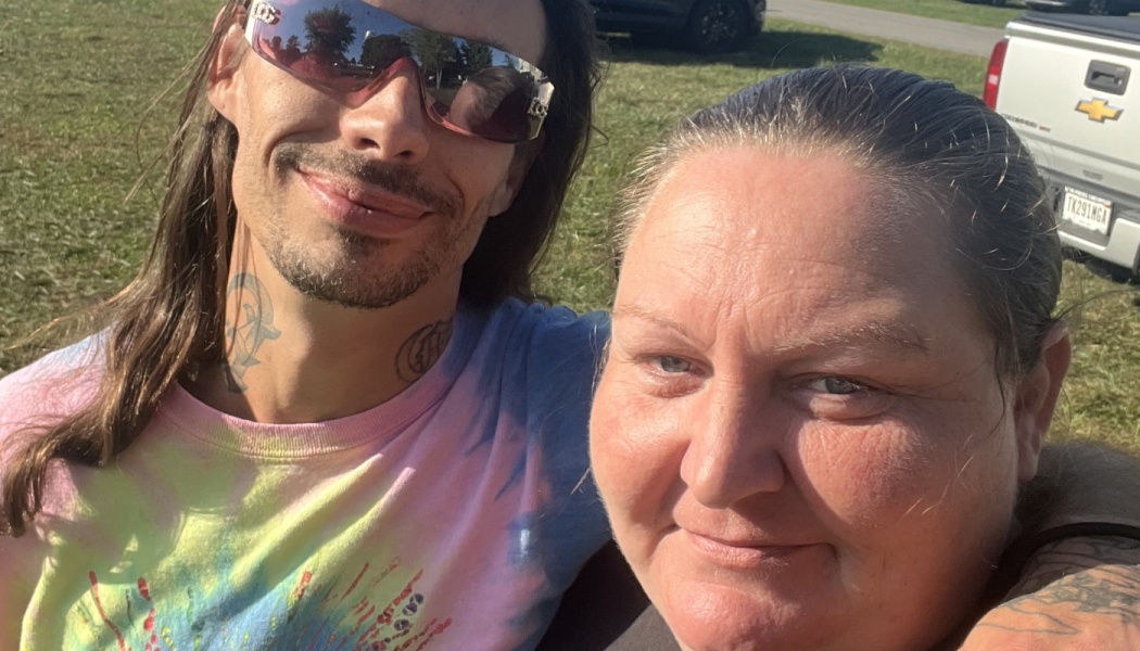 A smiling man with long hair, sunglasses, and a tie-dye shirt poses closely with a woman outdoors.