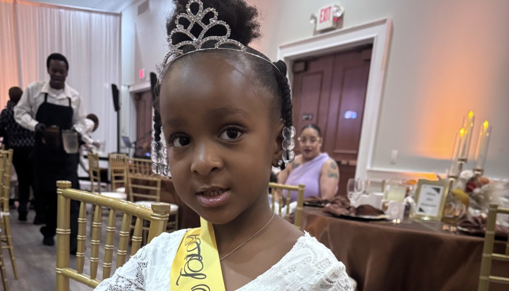 Young Black girl wearing a tiara, white dress, and yellow sash at a formal banquet event.