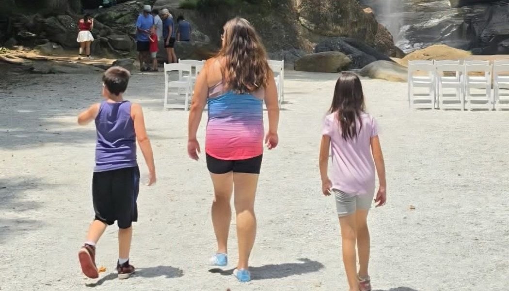 A mother and her two children walk across a sandy area toward rows of white folding chairs outdoors.