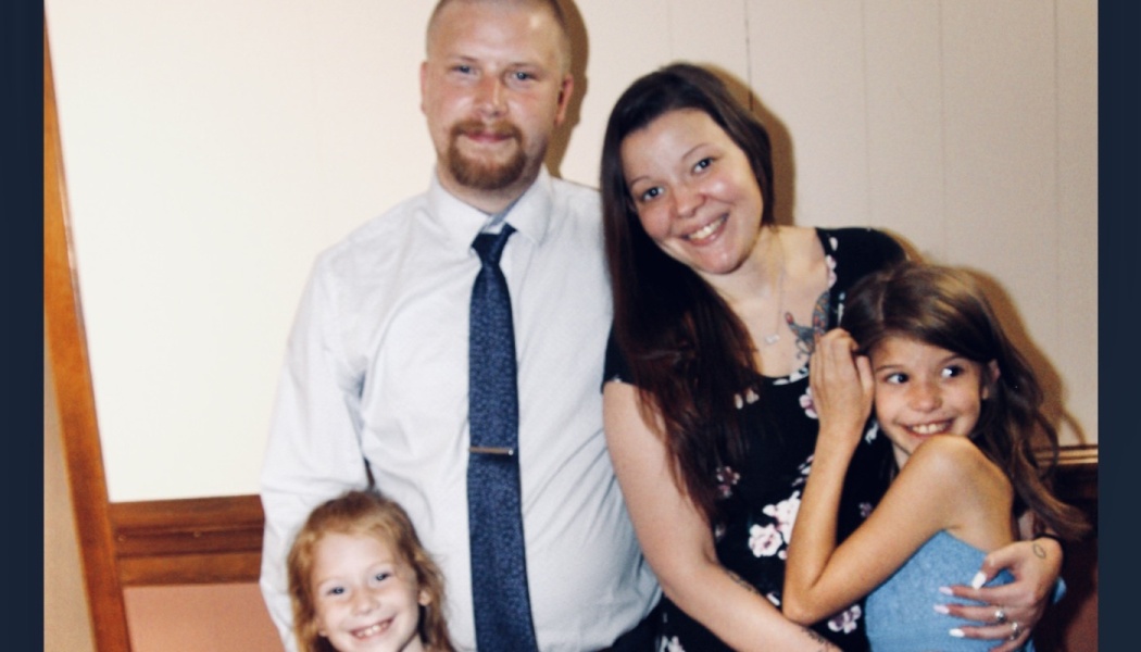 A smiling family of four posing together, with a father in a tie, a mother, and two young daughters.