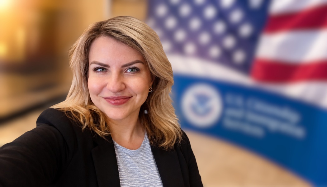 Smiling blonde woman in a black blazer with an American flag and USCIS seal blurred in the background.