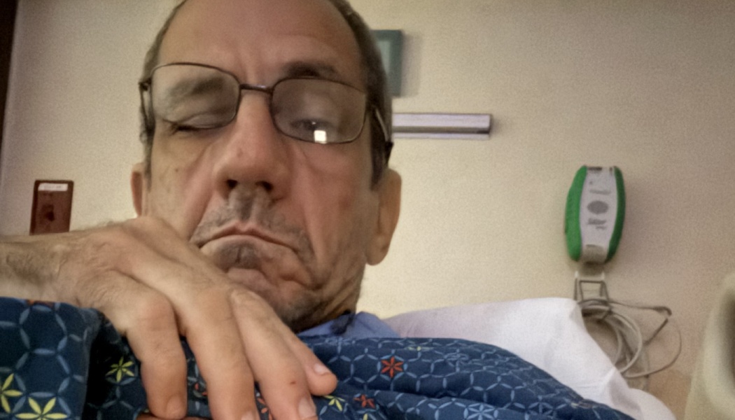 A man with glasses rests in a hospital bed wearing a blue patterned hospital gown.