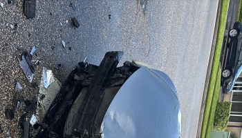 Heavily damaged front end of a silver car with debris scattered on asphalt after a crash.