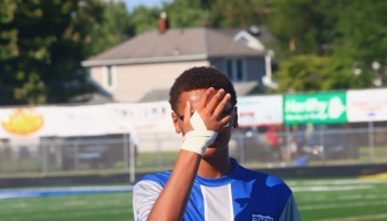 Young athlete in a blue sports jersey covers his face with his bandaged hand on an outdoor field.