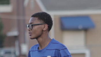 A young Black man wearing glasses and a blue striped shirt, looking thoughtfully to the side outdoors.