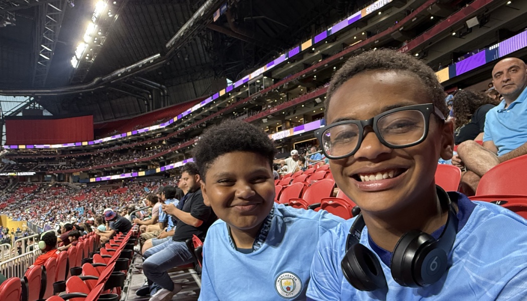 Two smiling boys in light blue jerseys pose for a selfie in the stands of a crowded stadium.