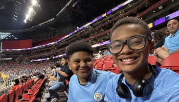 Two smiling boys in light blue jerseys pose for a selfie in the stands of a crowded stadium.