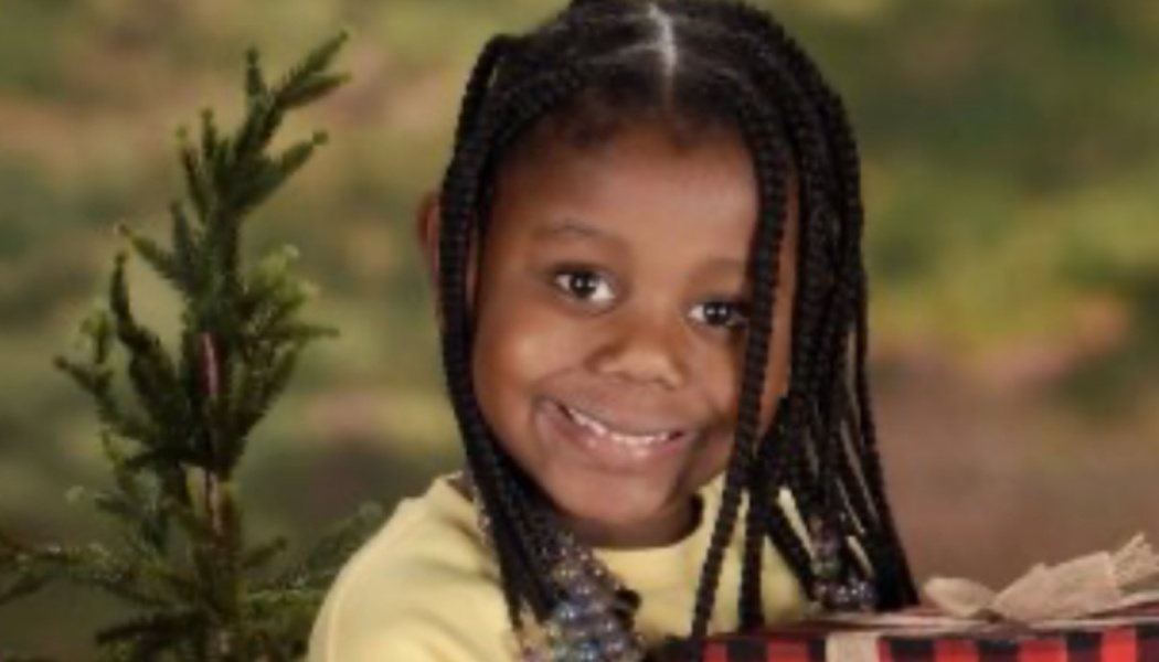 Smiling young girl with braided hair holding a wrapped Christmas gift next to a small pine tree.