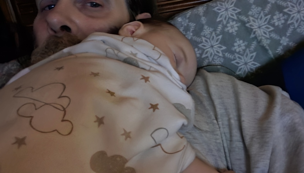 A man lies in bed with a sleeping baby wrapped in a star-patterned blanket resting on his chest.