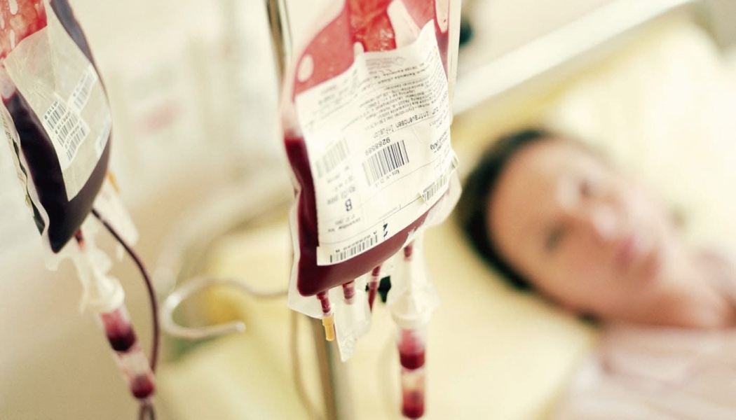 Blood transfusion bags hanging on an IV pole with a blurred patient lying in a hospital bed in the background.