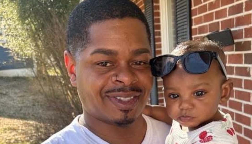 A smiling Black man holding a toddler wearing sunglasses on top of her head in front of a brick wall.