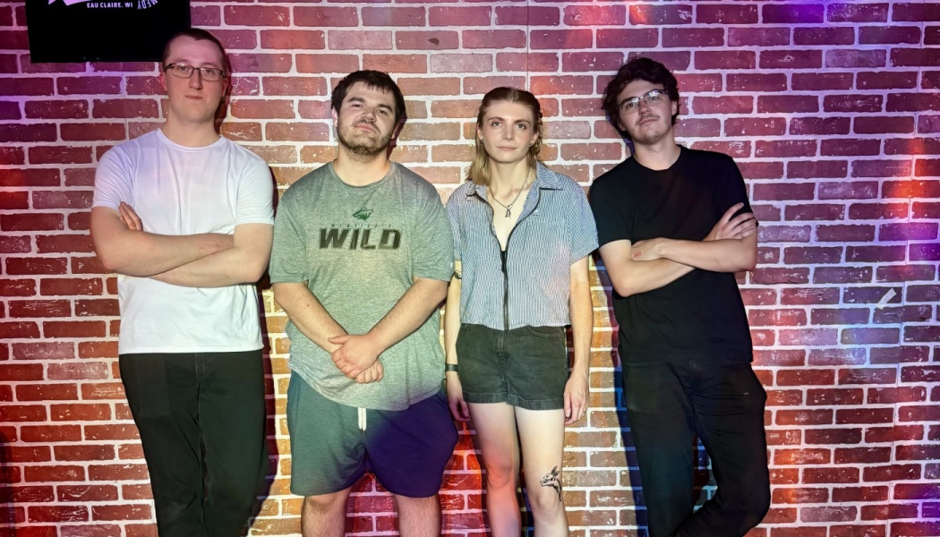 Four people standing side by side in casual clothing in front of an exposed red brick wall.