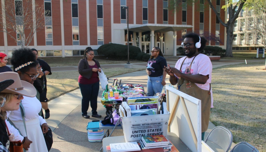 A smiling artist in an apron and headphones interacts with visitors at an outdoor college campus art booth.