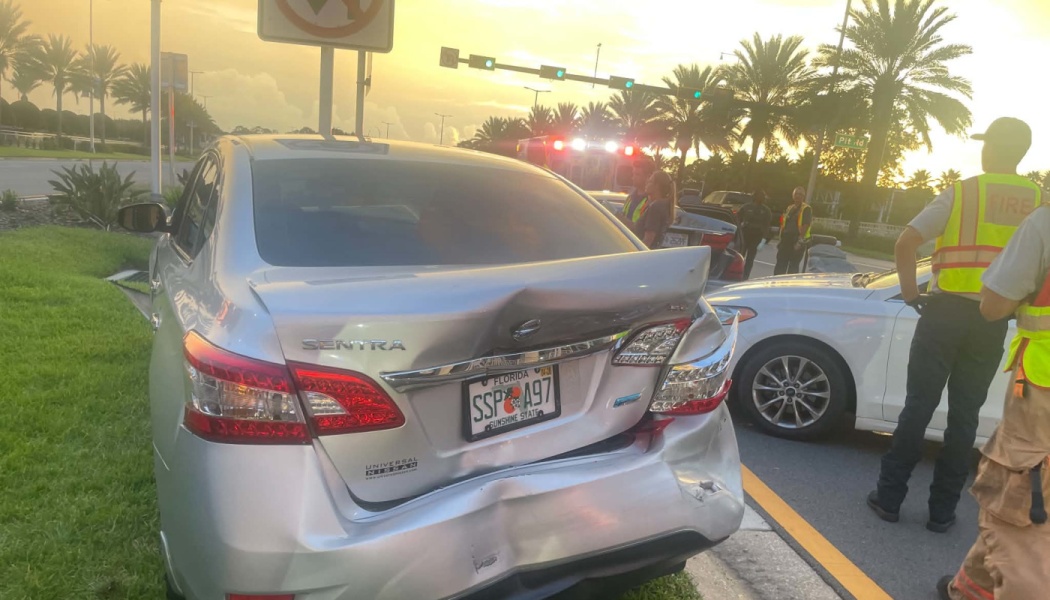 Silver Nissan Sentra with severe rear-end damage at a car crash scene with emergency responders.