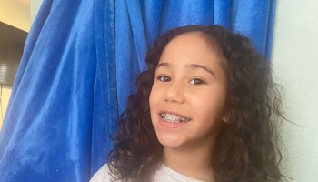 Young girl with long curly dark hair smiling and showing her braces in front of a blue curtain.
