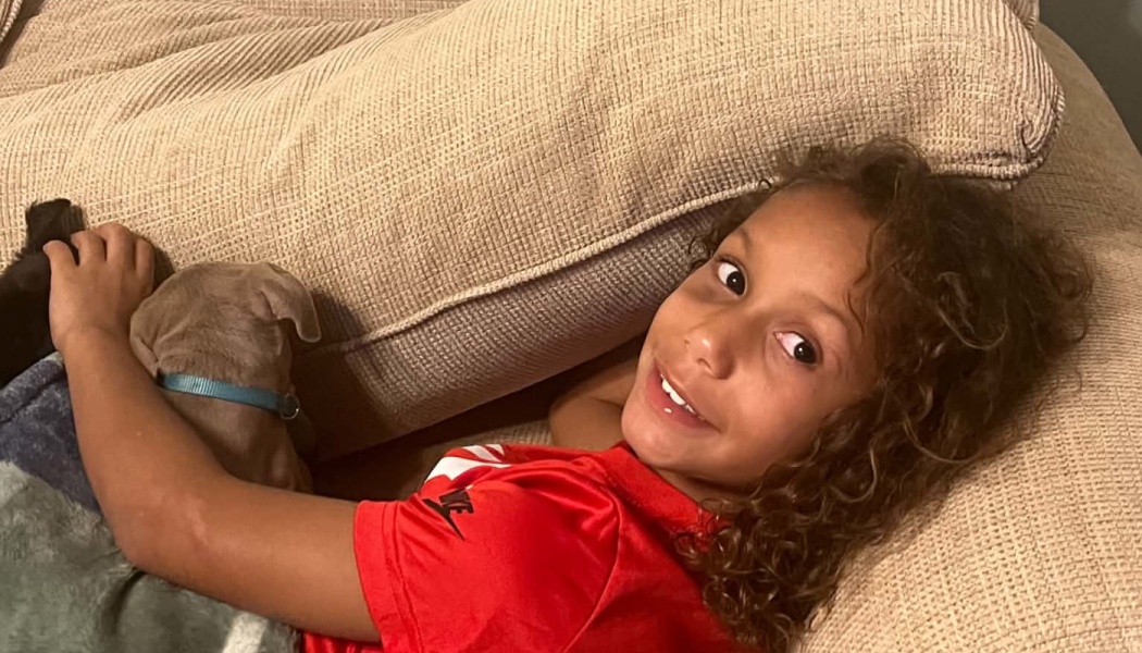 A smiling young boy with curly hair lies on a couch, cuddling a small puppy with a blue collar.