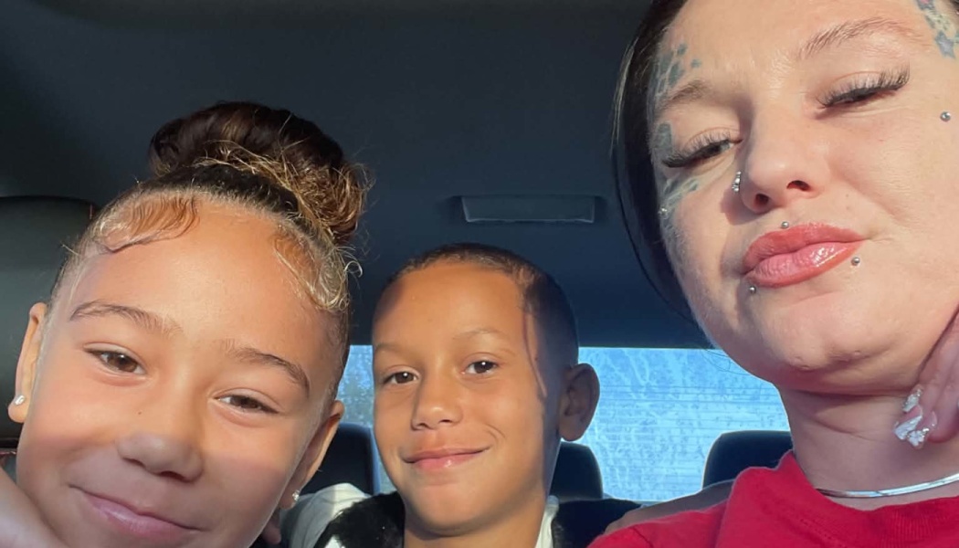 A mother and her two young children smiling together for a close-up selfie inside a car.