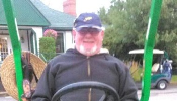 Smiling older man with a white goatee wearing a blue cap and black jacket, driving a green vehicle outdoors.