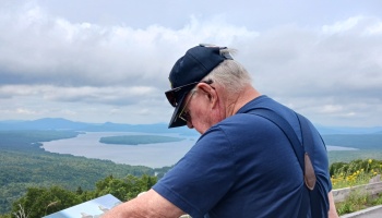 An elderly man wearing suspenders and a cap looks at a display board overlooking a scenic lake and mountains.