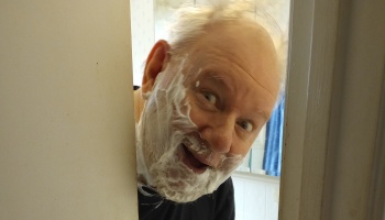 An older man with shaving cream on his face peeks cheerfully around a doorway.