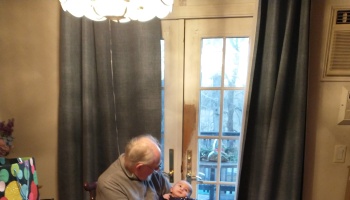An elderly man with glasses lovingly holding a baby while sitting near a window.