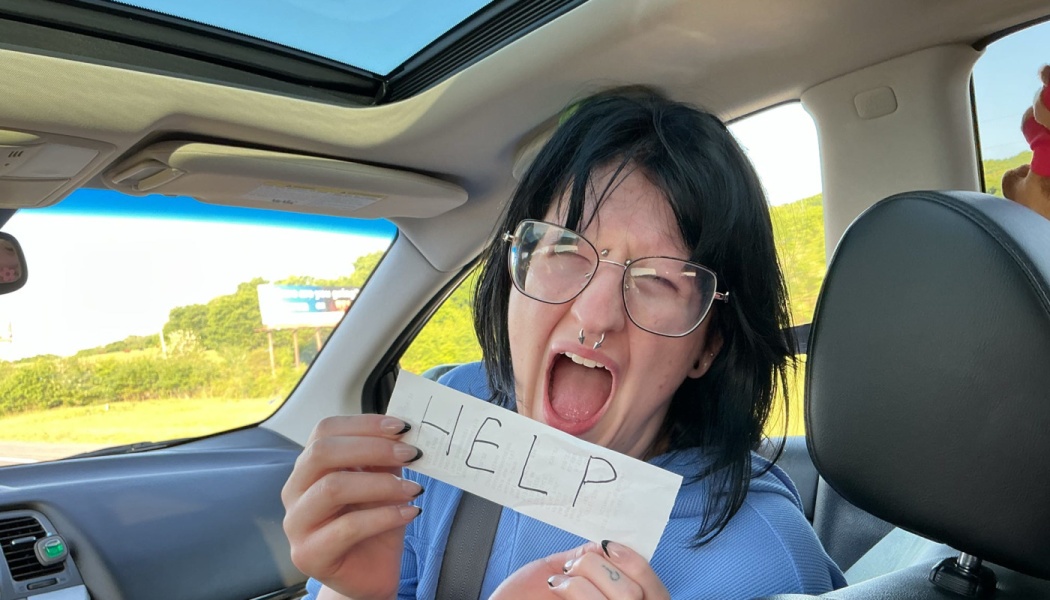A person with glasses and dark hair screaming in a car while holding a slip of paper that reads HELP.