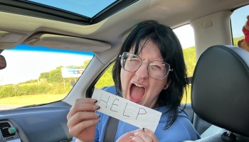 A person with glasses and dark hair screaming in a car while holding a slip of paper that reads HELP.