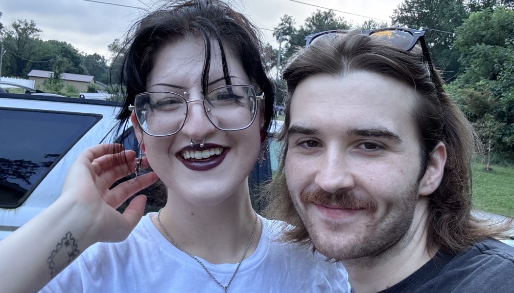 A smiling couple takes a close-up selfie outdoors. One wears glasses and dark lipstick; the other has a mustache.