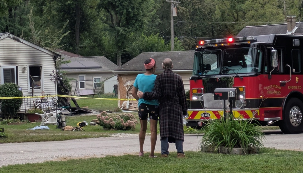 Two people embrace outside their fire-damaged house beside a parked red fire truck.