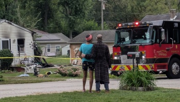 Two people embrace outside their fire-damaged house beside a parked red fire truck.