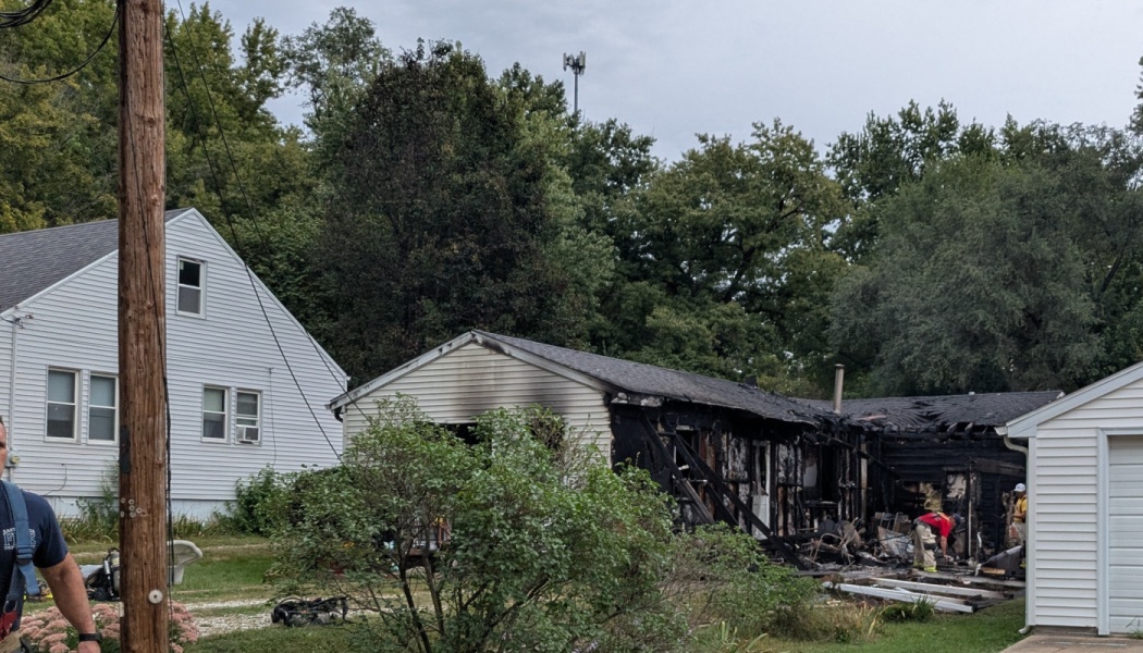 A house severely damaged by fire with charred walls, a collapsed roof, and firefighters on scene.