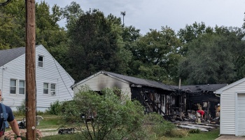 A house severely damaged by fire with charred walls, a collapsed roof, and firefighters on scene.