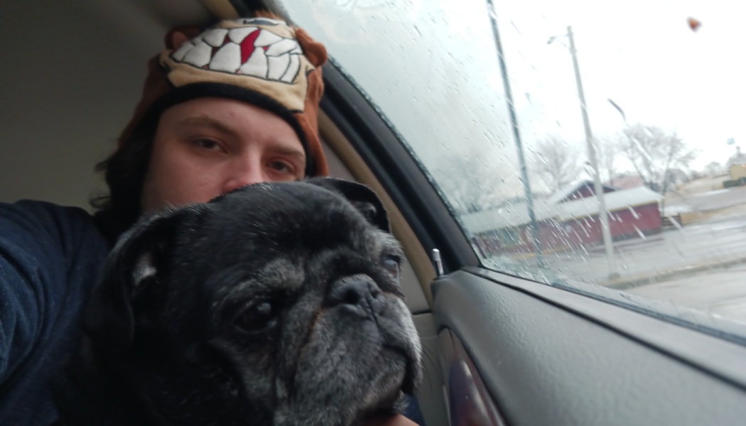A person wearing a cartoon beanie poses inside a car with a senior black pug on a rainy day.