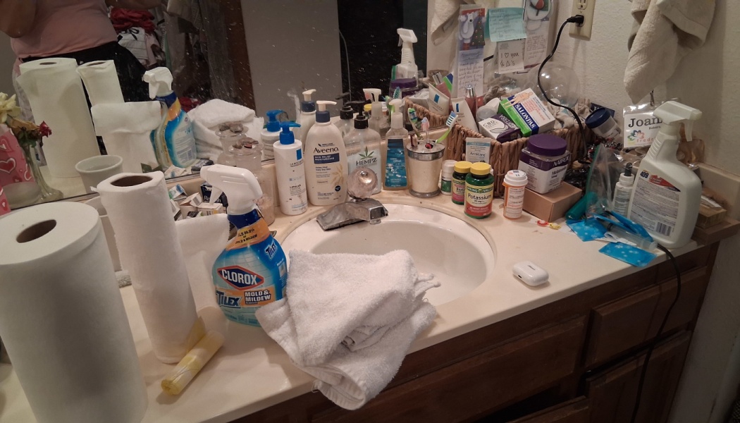 A bathroom counter cluttered with paper towel rolls, cleaning sprays, toiletries, and pill bottles around a white sink.