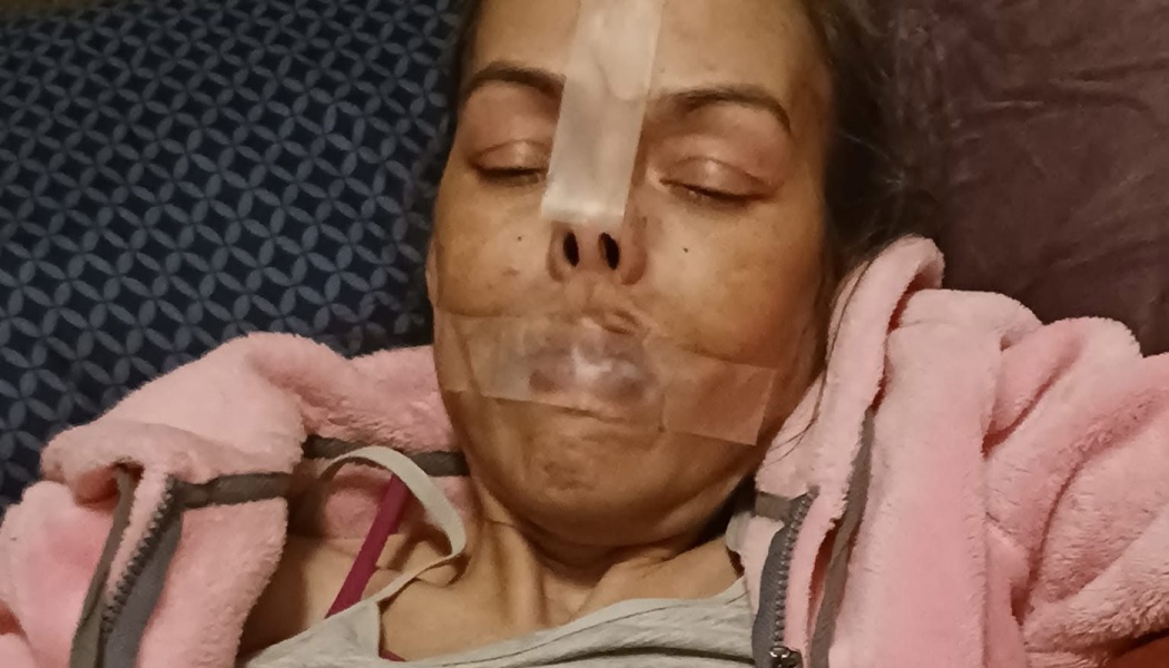 Woman resting in bed with medical tape across her nose and mouth, wearing a pink fleece jacket.