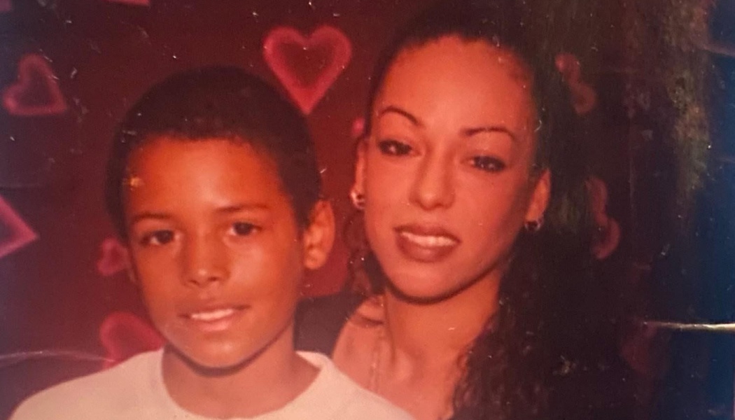 A young boy and a woman smiling together in front of a red heart-patterned background.