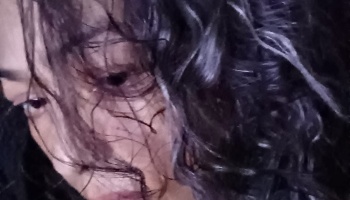 Close-up of a person's face partially obscured by dark, messy curls in dim light.