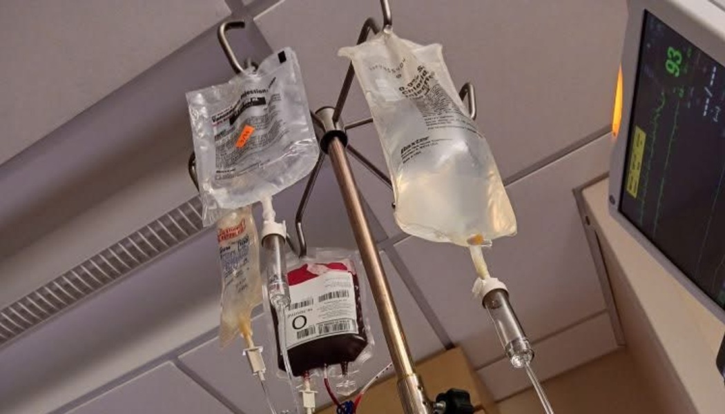 Medical IV pole holding clear fluid bags and a blood transfusion bag in a hospital room.