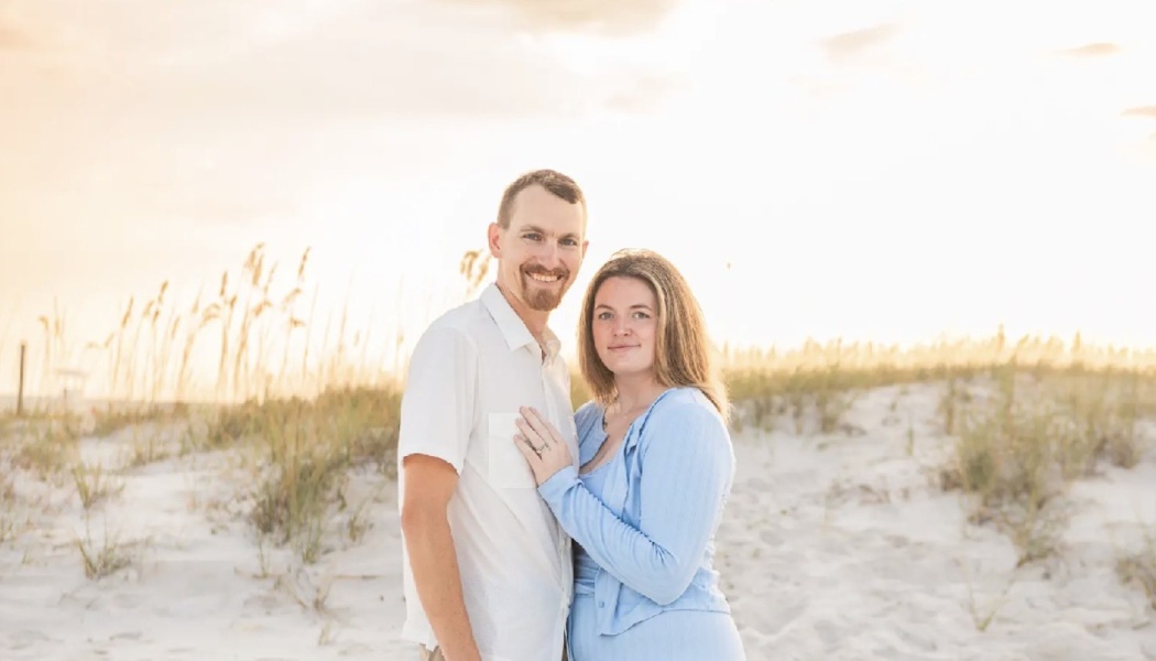 A smiling couple standing on a sandy beach dune at sunset, with the woman resting her hand on the man's chest.