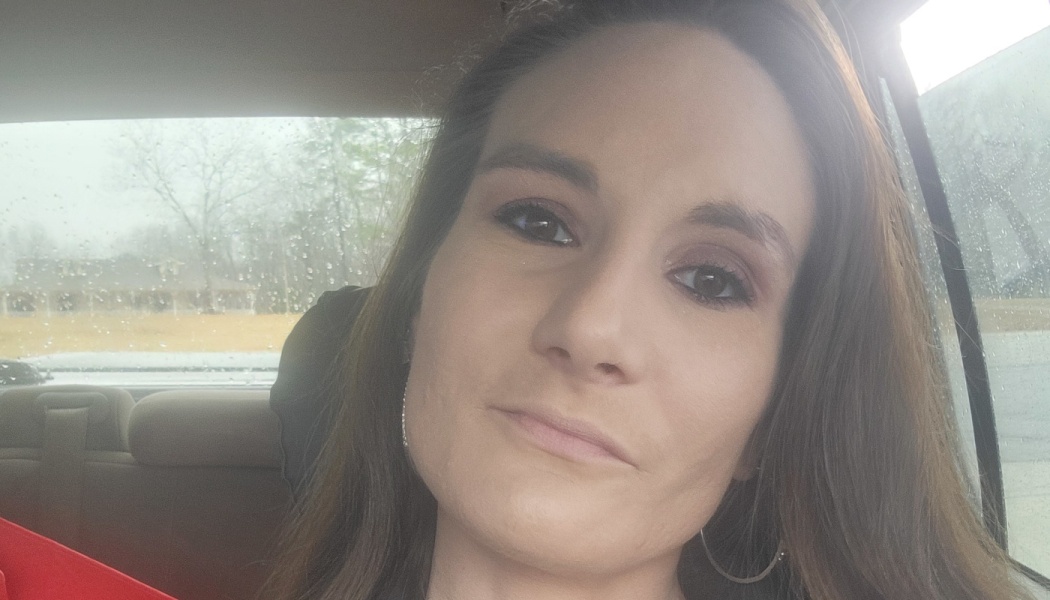 Close-up selfie of a woman with long brown hair and hoop earrings sitting inside a car on a rainy day.
