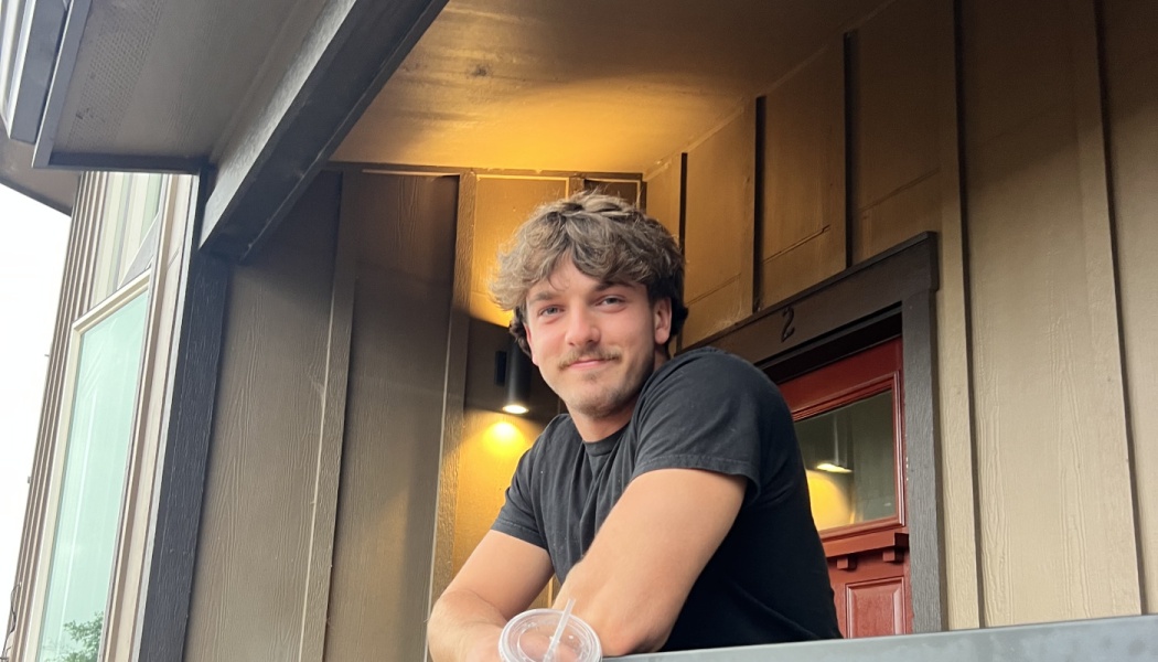 A smiling young man with wavy hair and a mustache leaning on a porch railing with an iced drink.