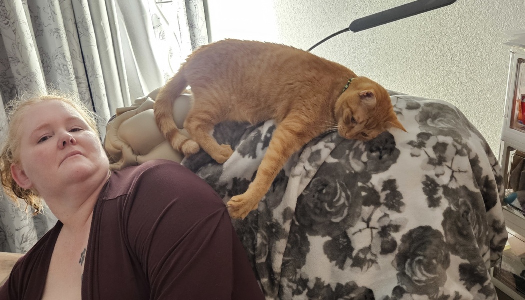 An orange tabby cat lies on a floral blanket behind a woman, resting its paw near her shoulder.